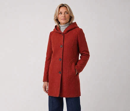 Frau in einem roten Kapuzenmantel und dunklen Jeans, steht mit der rechten Hand in der Manteltasche und blickt nach rechts.