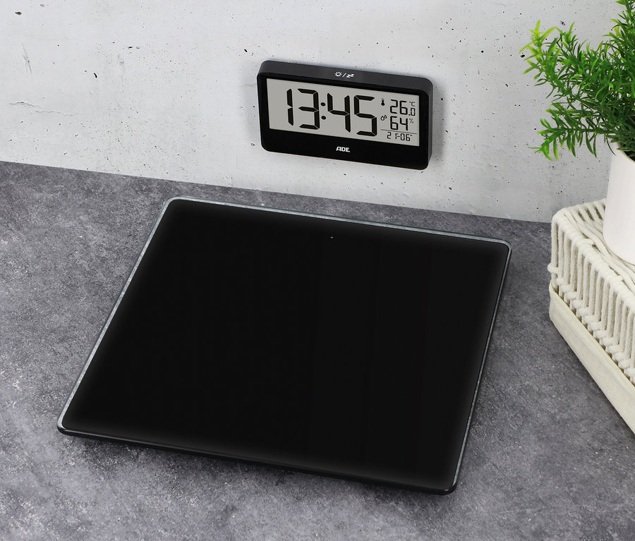 Eine schwarze digitale Personenwaage liegt auf einer grauen Oberfläche. Darüber zeigt eine schwarze Digitaluhr 13:45 Uhr, 26.0°C, 64% Luftfeuchtigkeit und das Datum an. Rechts steht eine grüne Pflanze in einem weißen Topf auf einem geflochtenen Korb.