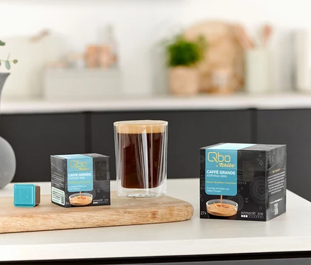 Auf einem Tisch stehen eine Tasse Kaffee, ein Qbo Caffè Grande KIRINYAGA BAYA - 8 Kapseln und ein Qbo Caffè Grande KIRINYAGA BAYA - 27 Kapseln.