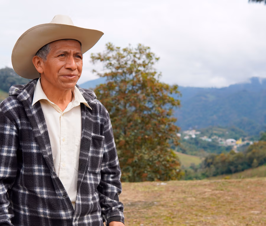 Ein älterer Mann mit hellem Cowboyhut und schwarz-weiß karierter Jacke steht im Freien und blickt nach rechts. Im Hintergrund sind grüne Berge, ein Tal mit einem Dorf und ein bewölkter Himmel zu sehen.