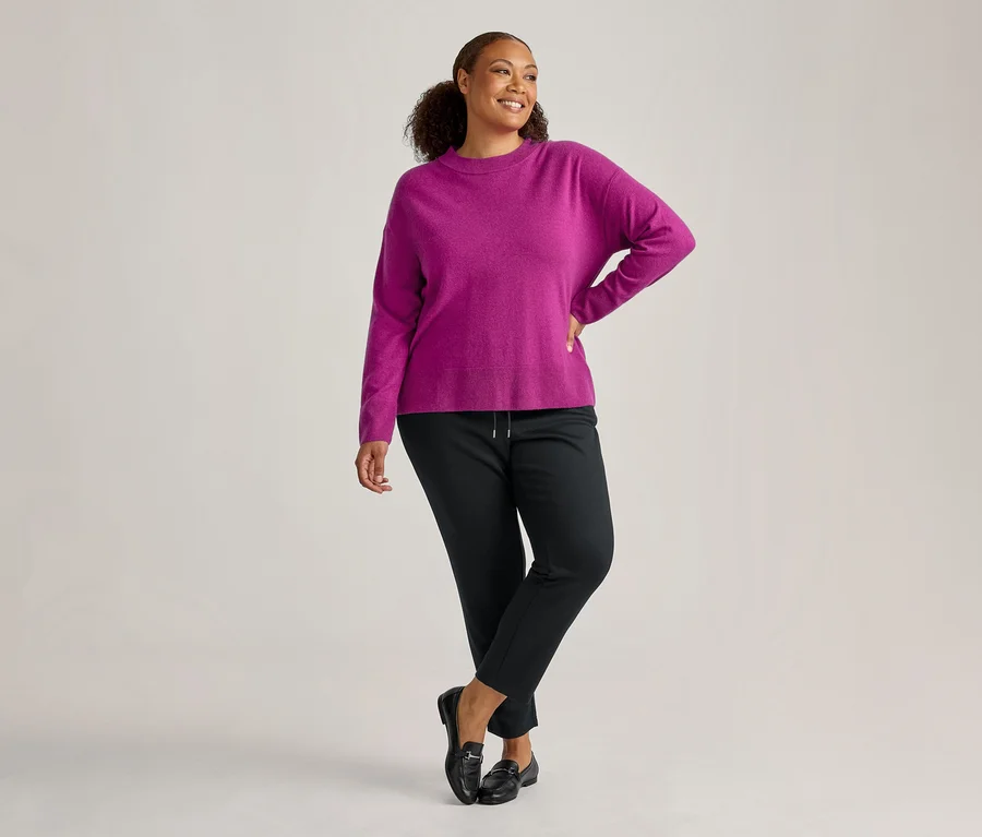 Lachende Frau in einem magenta Pullover, schwarzen Hosen und schwarzen Loafern, die mit der Hand in der Hüfte zur Seite blickt.