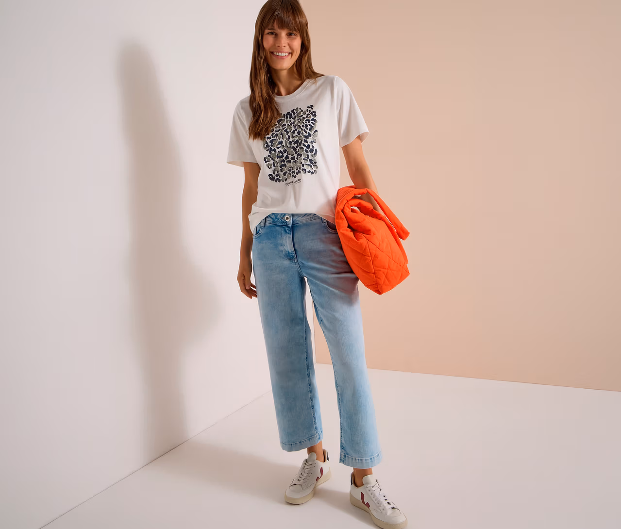 Eine lächelnde Frau steht in einem weißen CECIL Jerseyshirt mit Leopardenmuster, hellblauen Jeans und weißen Sneakern, eine orangefarbene Stepptasche haltend.