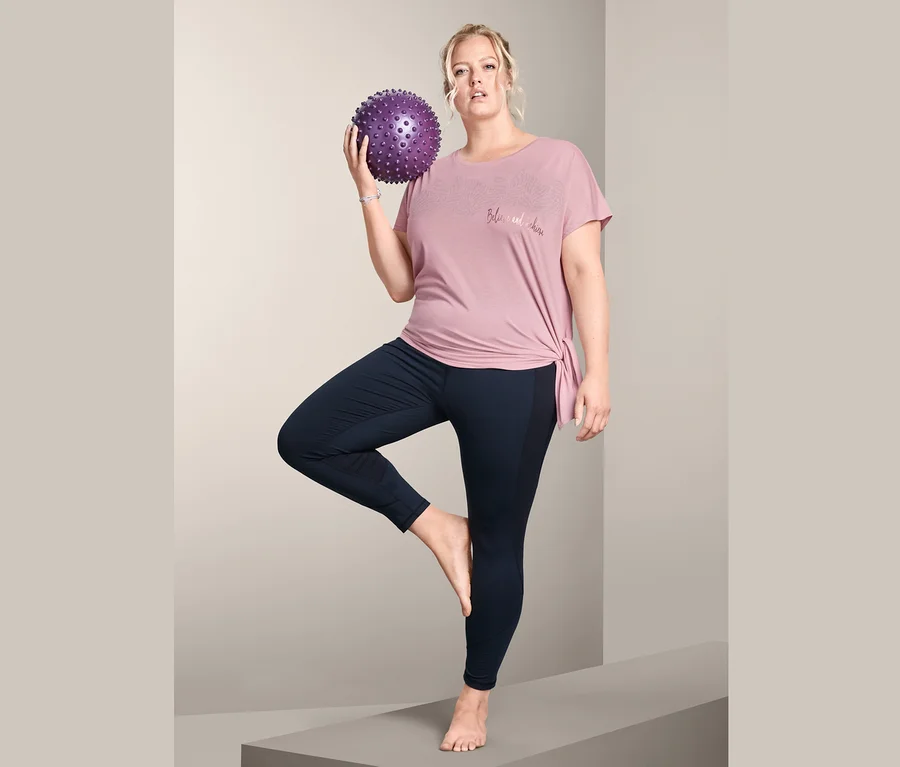 Frau balanciert auf einem Bein mit einem lila Massageball in der Hand, und trägt ein Sportshirt.