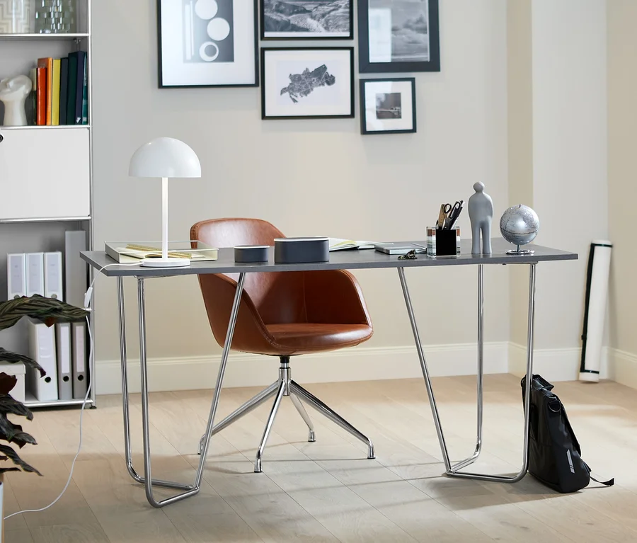 Innenraum mit Designtisch in Steinoptik mit Metall-Gestell, gepolstertem Drehstuhl aus Kunstleder und LED-Metall-Tischleuchte. Im Hintergrund befinden sich ein hohes Metallregal und ein Metall-Sideboard.
