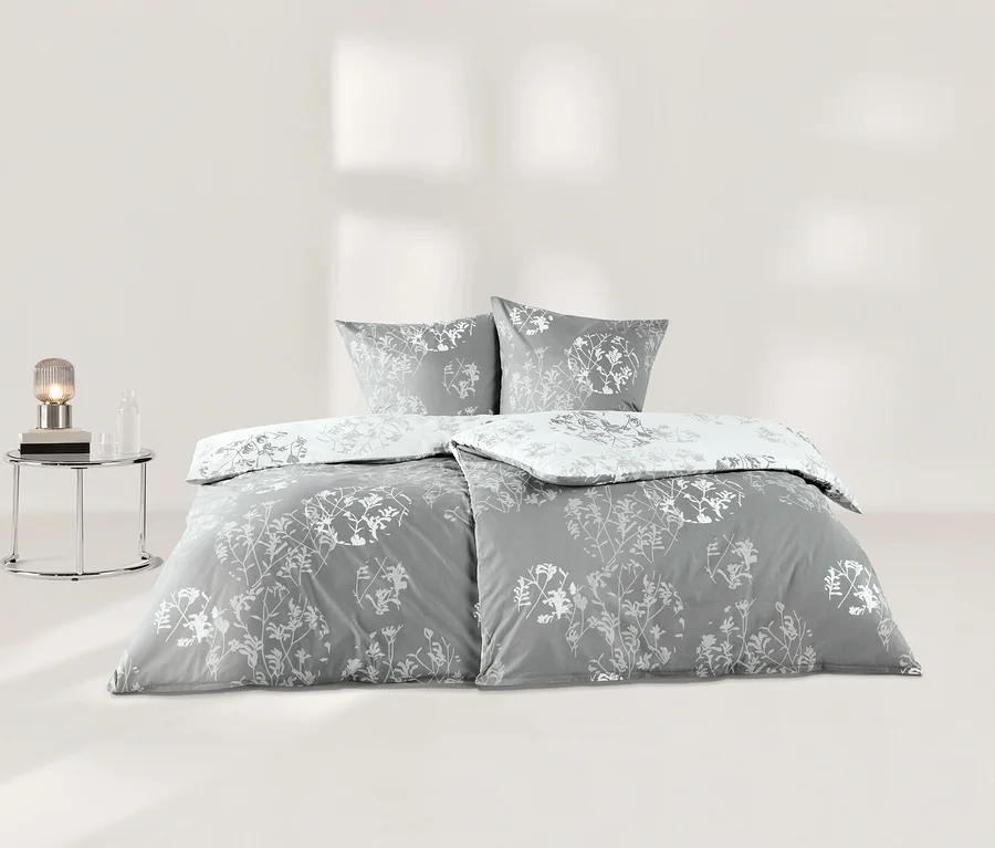 Graue Renforcé-Bettwäsche mit weißen Pflanzenmotiven auf einem Bett, bestehend aus zwei Kissen und einer Bettdecke. Links daneben steht ein Nachttisch mit einer Lampe und einer Flasche.