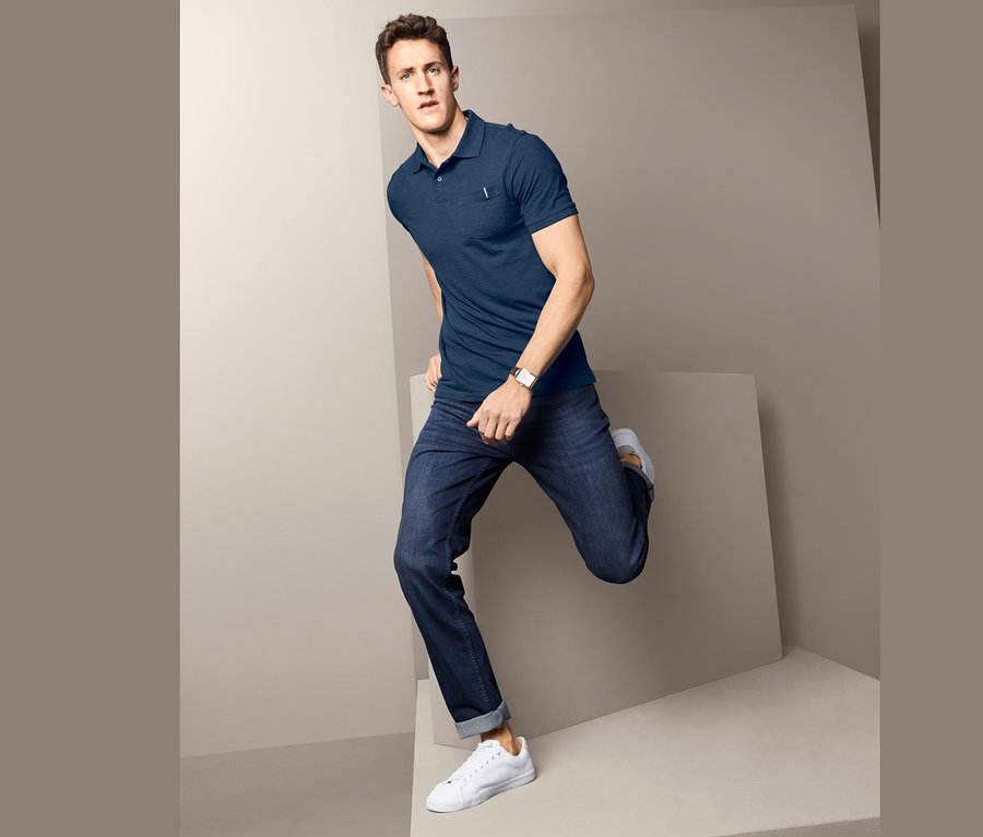 Mann posiert in blauem Poloshirt mit Strukturgarn, Jeans und weißen Schuhen vor grauem Hintergrund.