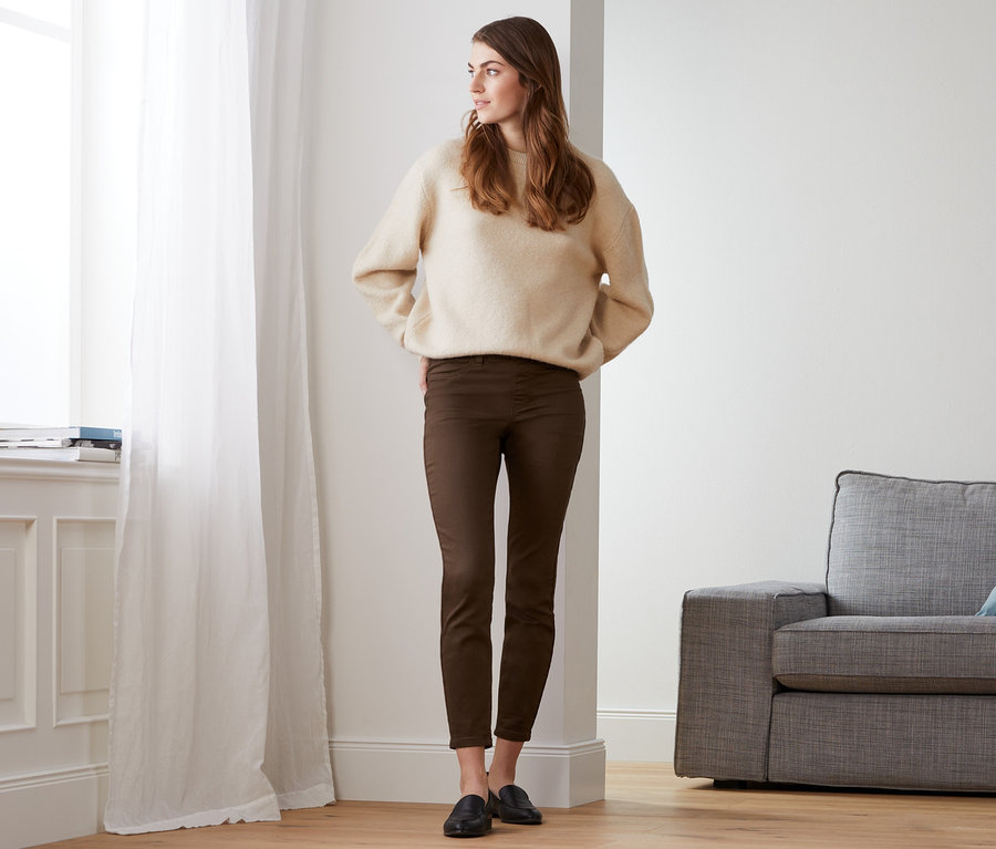 Frau steht im Raum am Fenster und trägt einen beigen Pullover und Treggings.