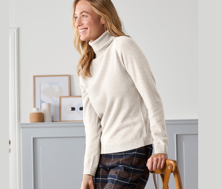 Eine lächelnde Frau lehnt an einem Stuhl und trägt einen Cashmere-Feinstrickpullover und eine karierte Hose.
