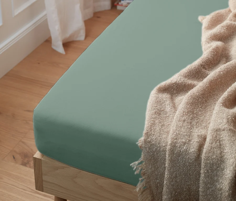 Grünes Jersey-Spannbettlaken auf einem Bett mit einer beigen Decke.
