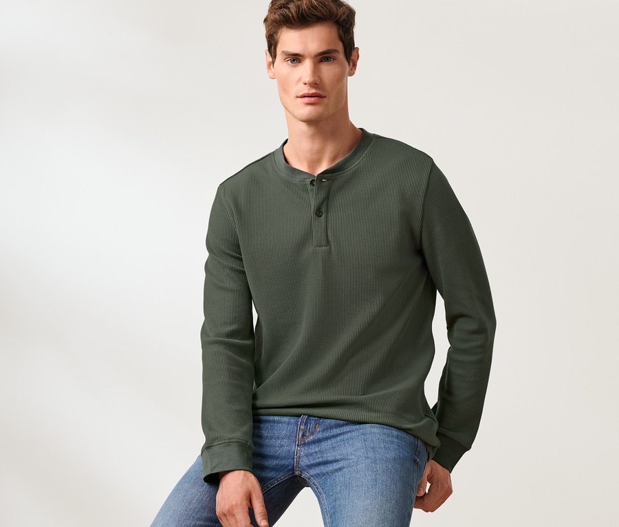 Mann sitzt auf einem Hocker und trägt ein Langarm-Henley und Jeans.