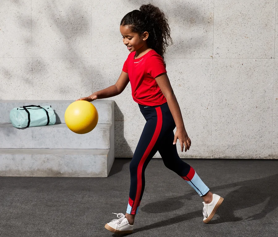 Mädchen dribbelt in rotem T-Shirt mit gelbem Ball. Sie trägt eine Highwaist-Sporttight mit recyceltem Polyester, colorblocking.