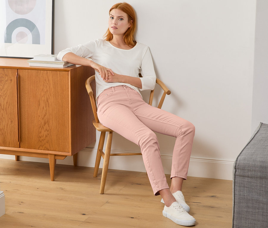 Eine Frau sitzt auf einem Stuhl und trägt eine roséfarbene Slimfit-Jeans.