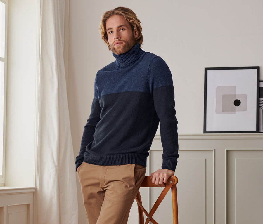 Mann, der sich an einen Stuhl lehnt, trägt einen Rollkragen-Pullover, braune Hose und weiße Turnschuhe.