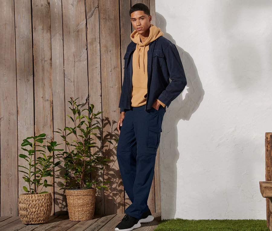 Mann steht und trägt eine marineblaue 2-in-1-Funktionshose, einen gelben Hoodie und eine Jacke.
