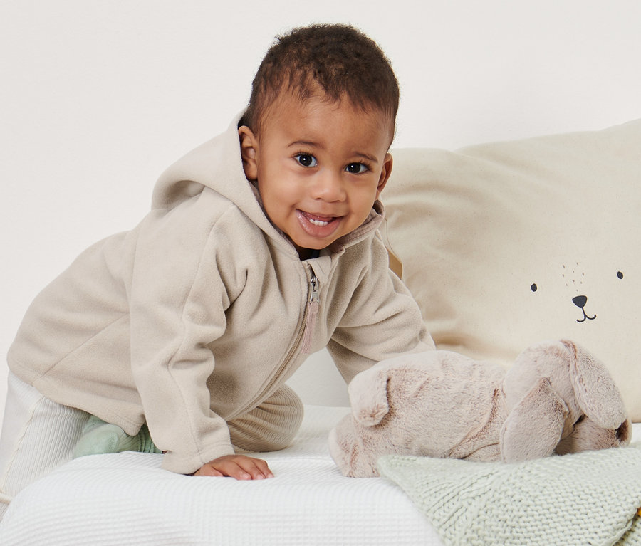 Ein lächelndes Baby krabbelt auf einem weißen Bett und trägt eine Baby-Fleecejacke.