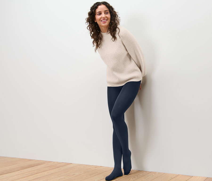 Lächelnde Frau, die sich an eine Wand lehnt, trägt eine Thermostrumpfhose und einen Pullover.