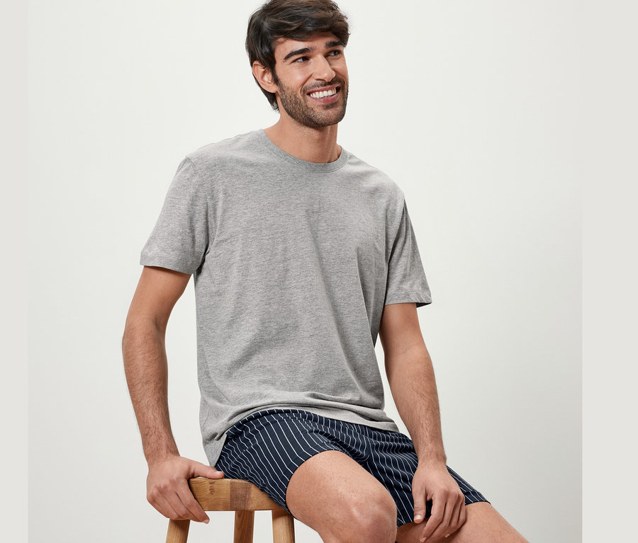Mann sitzt auf einem Hocker und trägt ein graues Shorty-Pyjama-Set.
