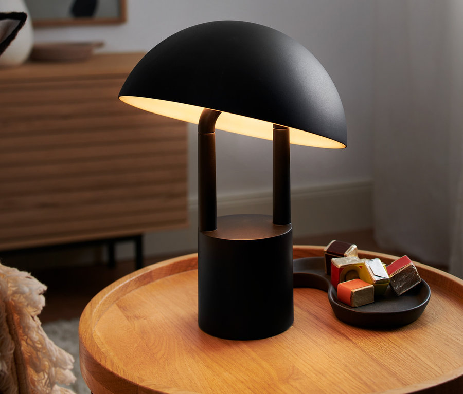 Eine schwarze LED-Akku-Tischleuchte steht auf einem Holztisch neben einem Tablett mit kleinen Päckchen.