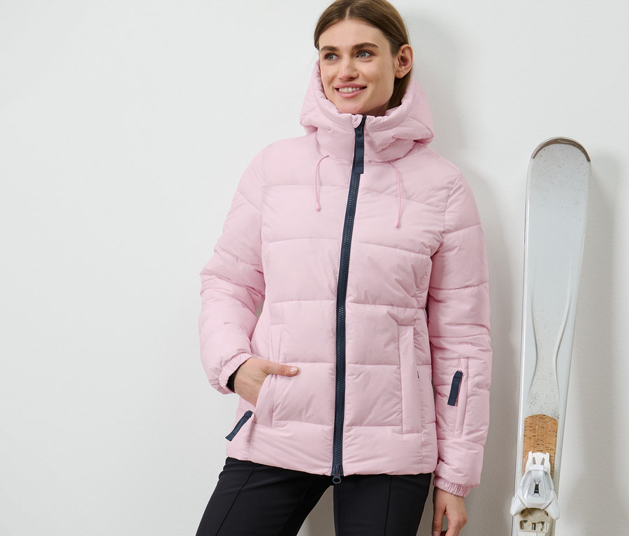 Frau steht neben Skiern und trägt eine rosa Ski-Steppjacke.
