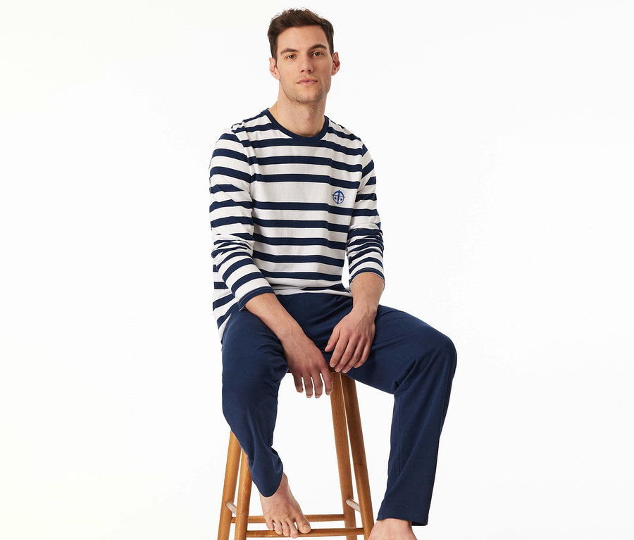 Mann sitzt barfuß auf einem Holzschemel und trägt ein SCHIESSER Pyjama-Set mit einem weiß-marineblau gestreiften Langarmshirt und einer marineblauen Hose.