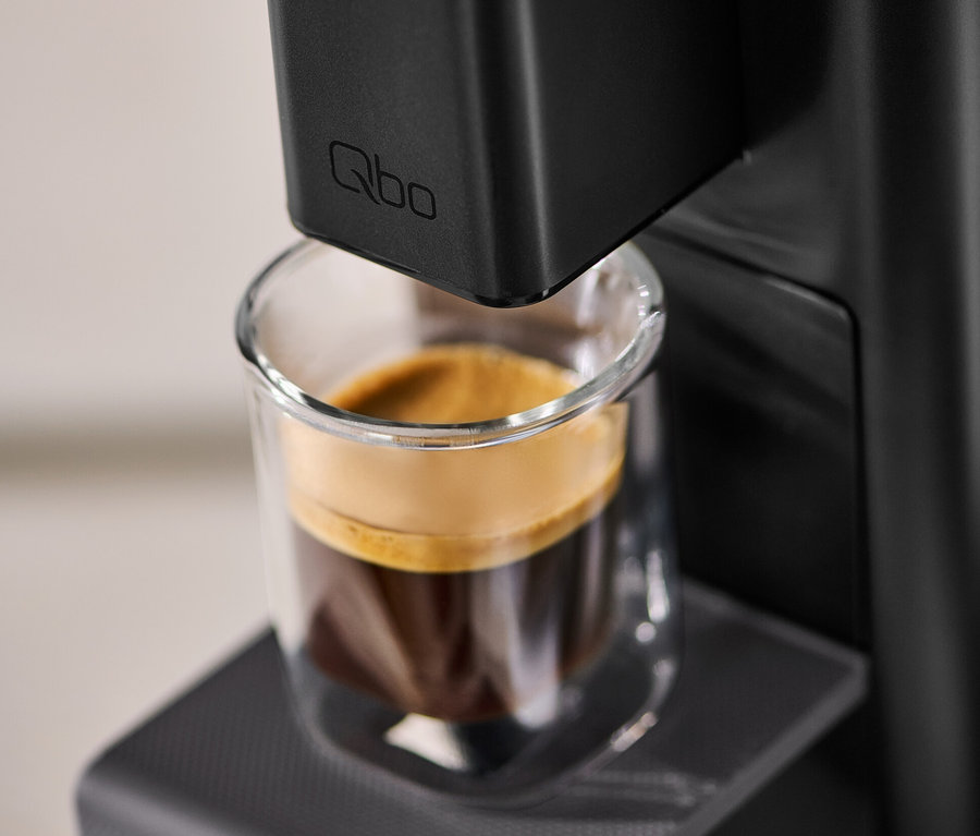 Nahaufnahme einer Qbo ICONIC Dark Chrome Kaffeemaschine mit einer Tasse Kaffee.