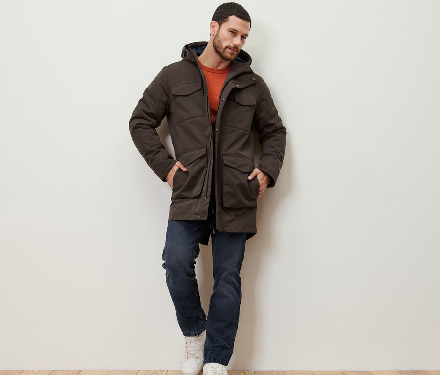 Mann lehnt an einer Wand und trägt eine Parka.