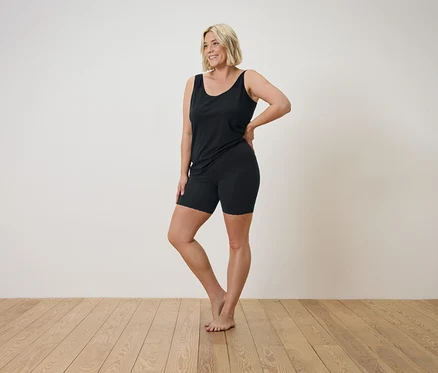 Blonde Frau in schwarzem Tanktop und Shorts posiert auf einem Holzboden.