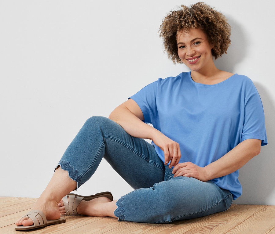 Frau sitzt auf Holzboden, trägt hellblaues Blusenshirt und 7/8-Treggings mit Lochstickerei.