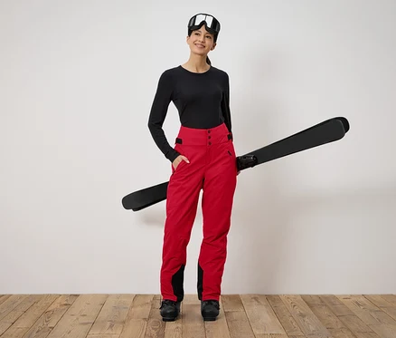 Frau steht und hält Ski. Sie trägt ein schwarzes Langarmshirt, eine rote Hose und eine Skibrille.