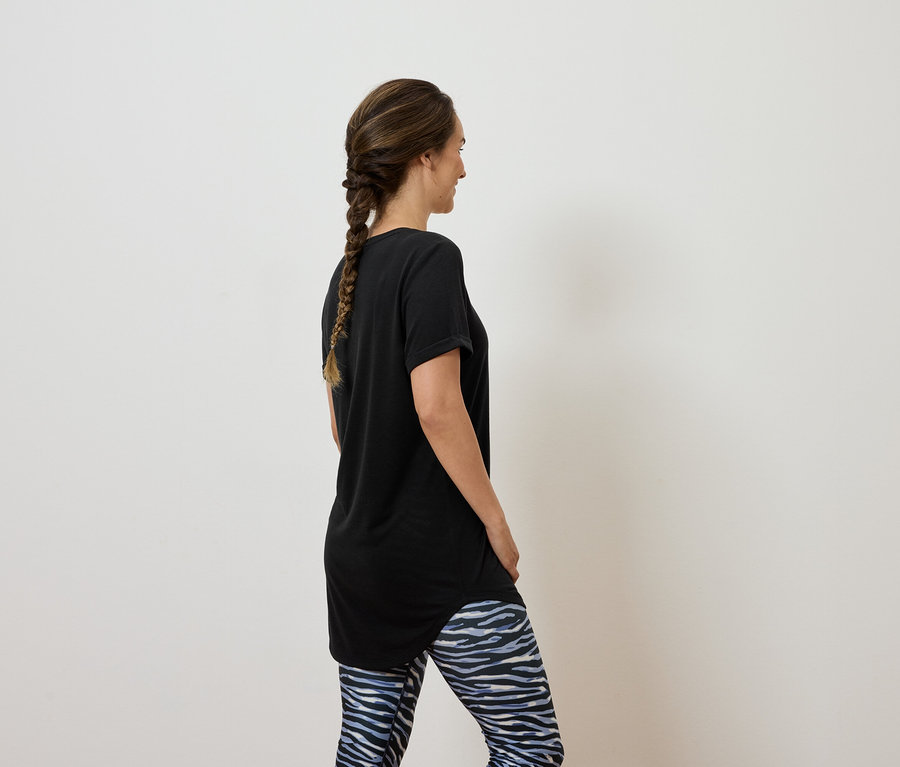 Frau mit braunen, zu einem Zopf geflochtenen Haaren steht mit dem Rücken zur Kamera und trägt ein schwarzes T-Shirt und blau-weisse Leggings.