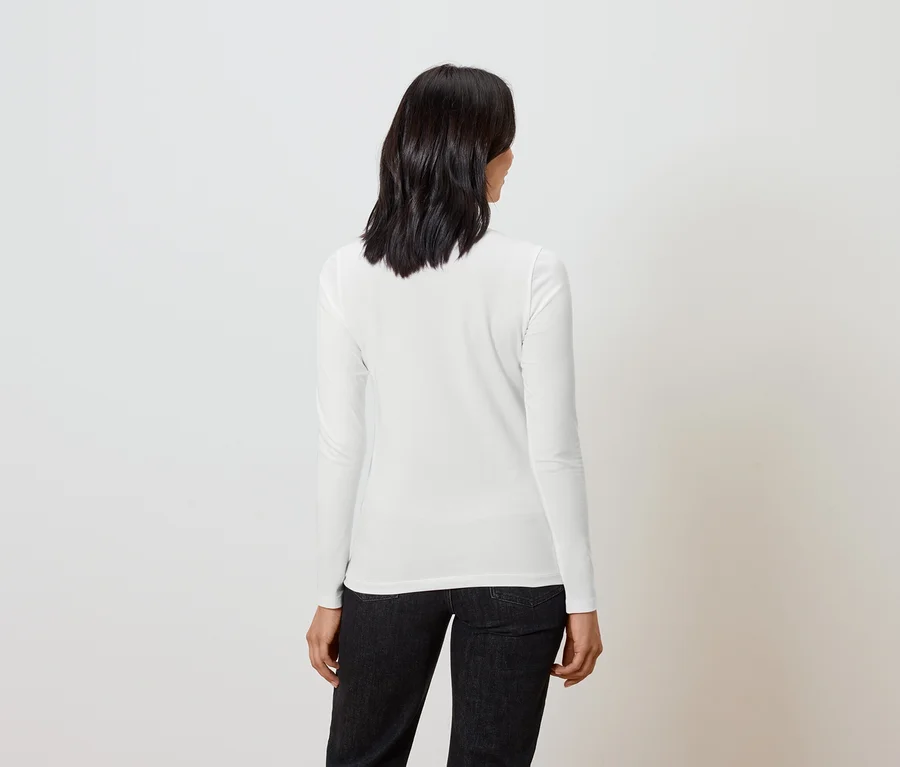 Frau mit kurzem dunklem Haar steht mit dem Rücken zur Kamera und trägt ein weißes Langarmshirt und schwarze Jeans.