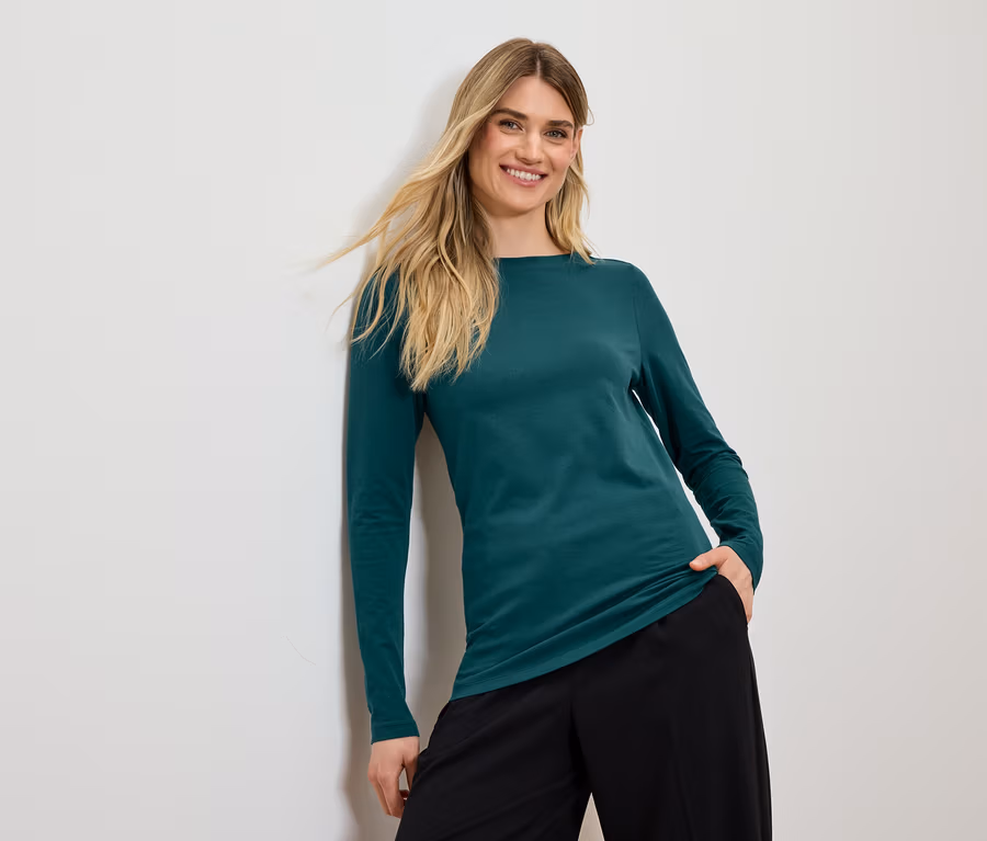 Lächelnde Frau mit blonden Haaren posiert in einem grünen Langarmshirt und einer schwarzen Hose.