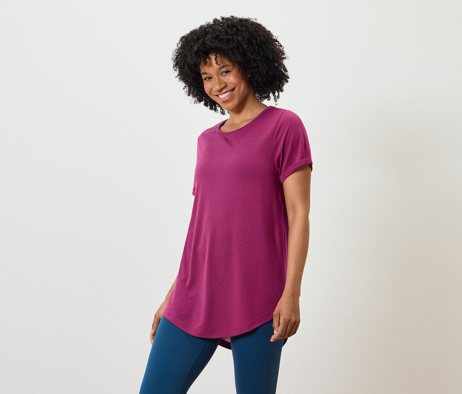 Frau mit dunklem lockigem Haar trägt ein purpurrotes T-Shirt und blaue Leggings.