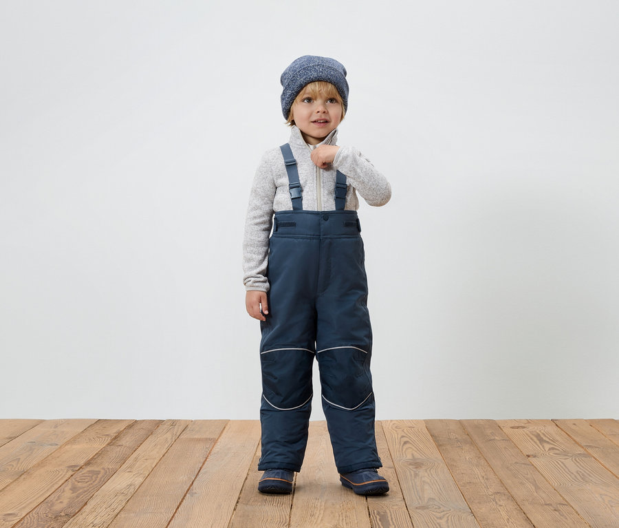 Ein Junge in blauer Winterkombination mit Hosenträgern und Strickmütze steht auf einem Holzboden und schließt seine Strickjacke.