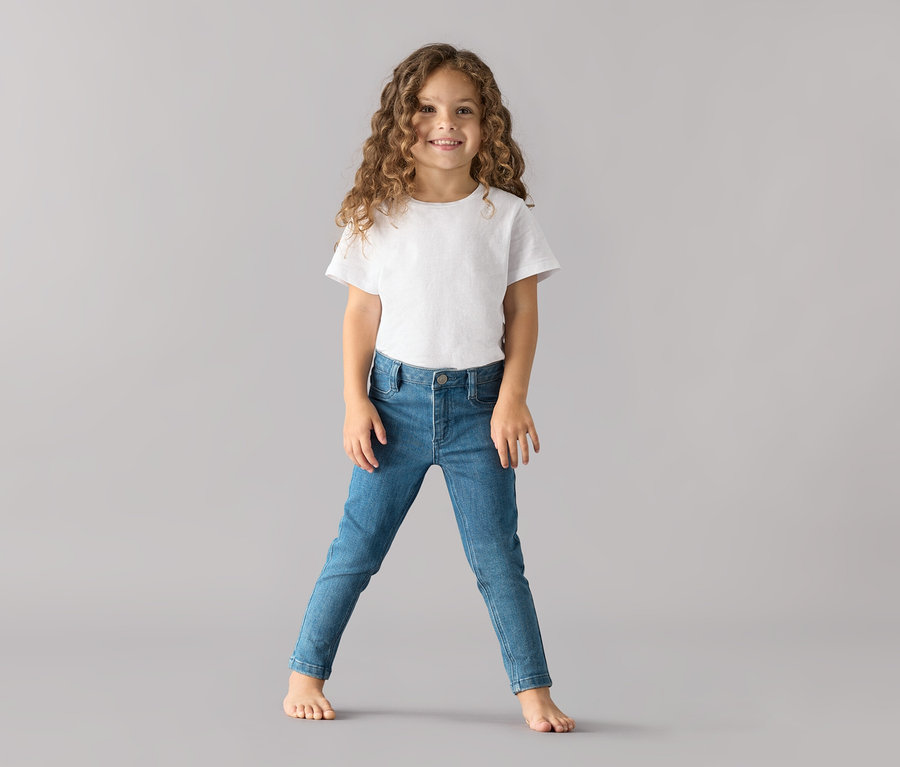 Mädchen in weißem T-Shirt und Kinder-Jeans – Fit »Lotta« steht vor grauem Hintergrund.