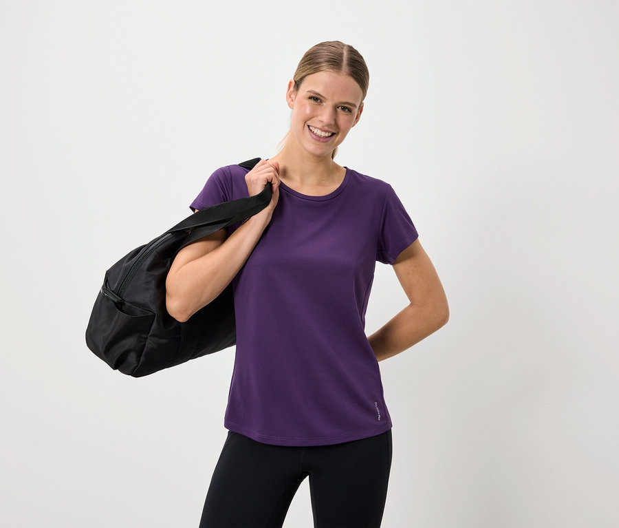 Lächelnde Frau in lila T-Shirt und schwarzen Leggings mit einer schwarzen Tasche über der Schulter.