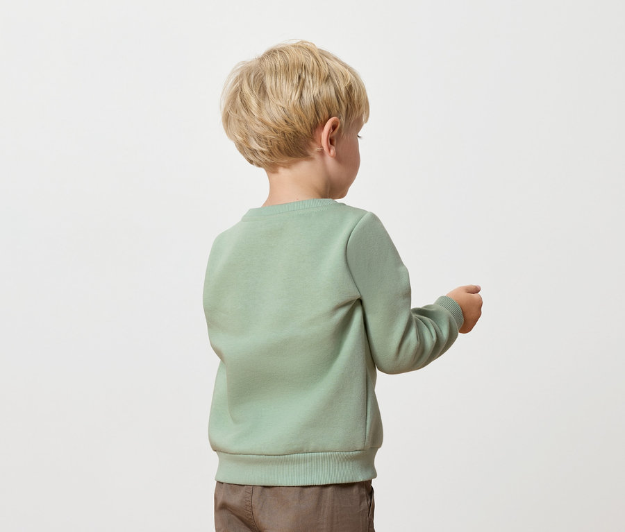 Ein blonder Junge, gekleidet in einem grünen Pullover und braunen Hosen, steht mit dem Rücken zur Kamera.