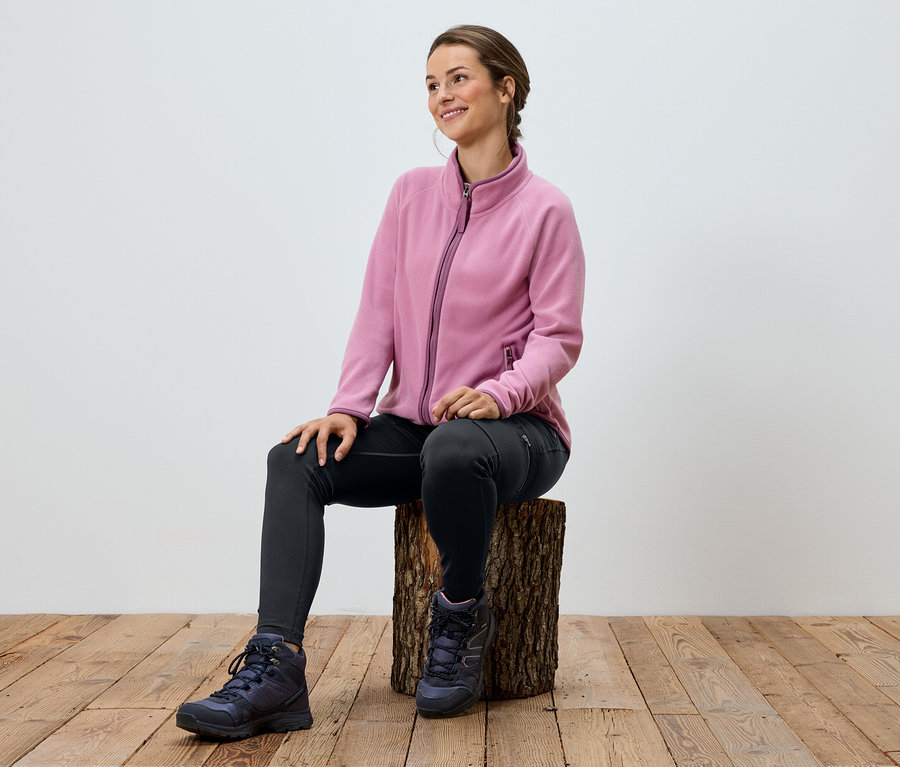 Frau sitzt auf einem Holzschemel, trägt eine rosa Fleecejacke, schwarze Leggings und Wanderschuhe.