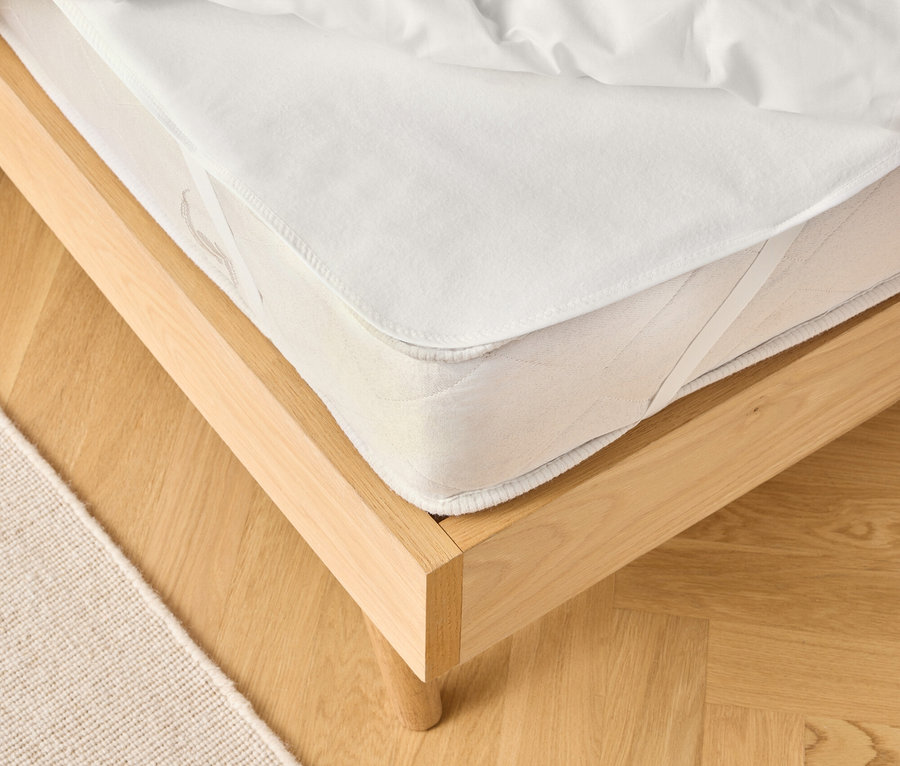 Ansicht eines hellen Holzbettsgestells mit einer weißen Matratze und einer weißen Bettdecke. Ein beiger Teppich ist teilweise sichtbar.