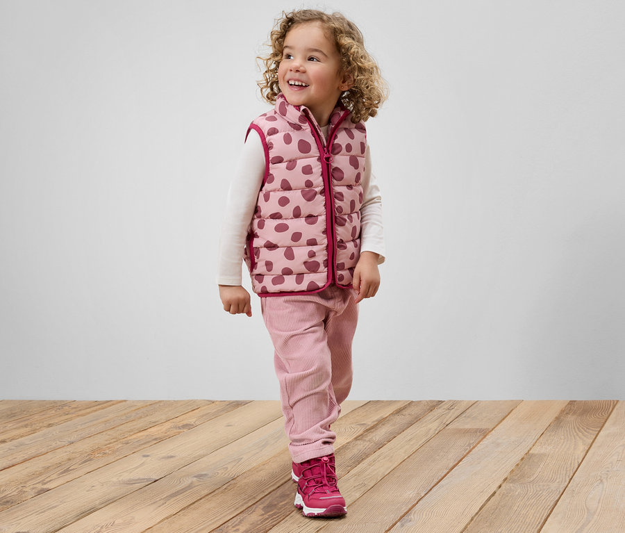 Ein lächelndes Mädchen mit lockigem Haar, gekleidet in eine rosa gepunktete Weste, rosa Hosen und rosa Schuhe, steht auf einem Holzboden.