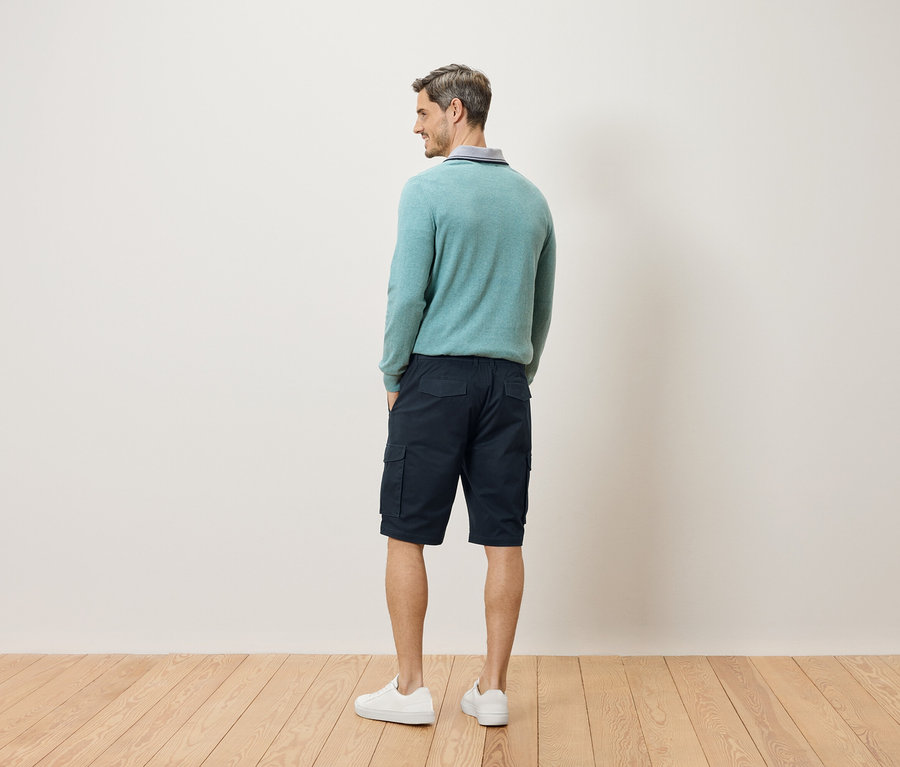 Ein Mann steht mit dem Rücken zur Kamera, trägt einen hellblauen Pullover, Cargoshorts und weiße Turnschuhe.