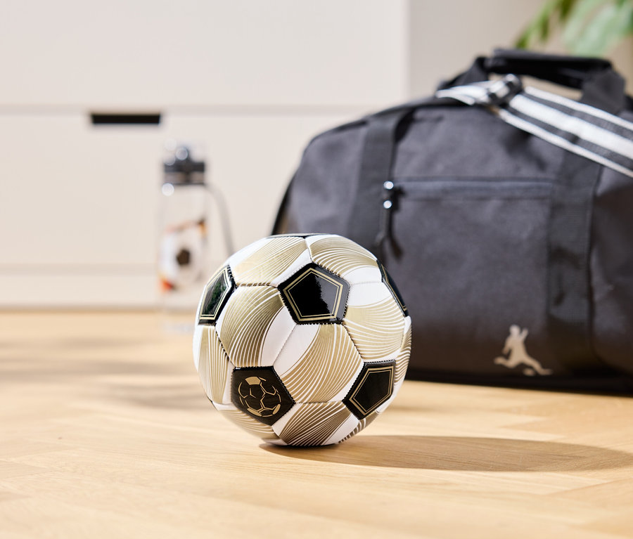 Ein goldfarbener Fußball liegt auf einem Holzboden, im Hintergrund eine Sporttasche und eine Wasserflasche.