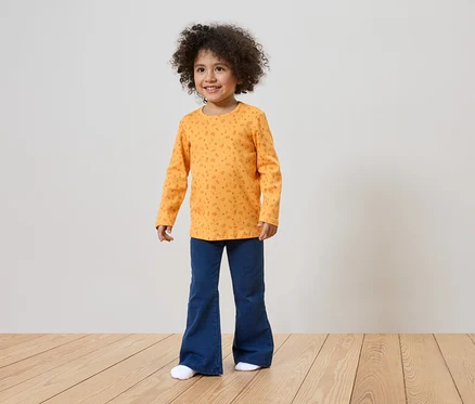 Ein lächelndes Mädchen mit lockigem Haar trägt ein orangefarbenes Langarmshirt mit Muster und blaue Schlaghosen.