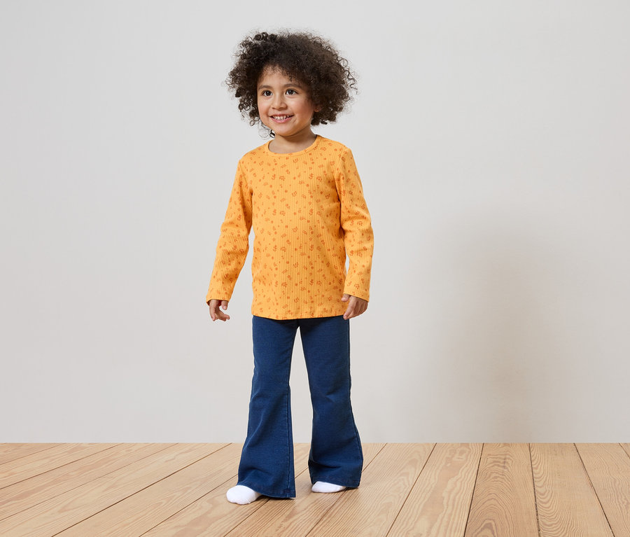 Ein lächelndes Mädchen mit lockigem Haar trägt ein orangefarbenes Langarmshirt mit Muster und blaue Schlaghosen.