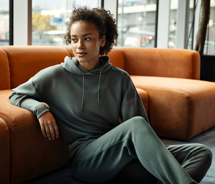 Frau sitzt auf dem Boden, an ein orangefarbenes Sofa gelehnt, und trägt einen grauen Hoodie und eine Jogginghose.