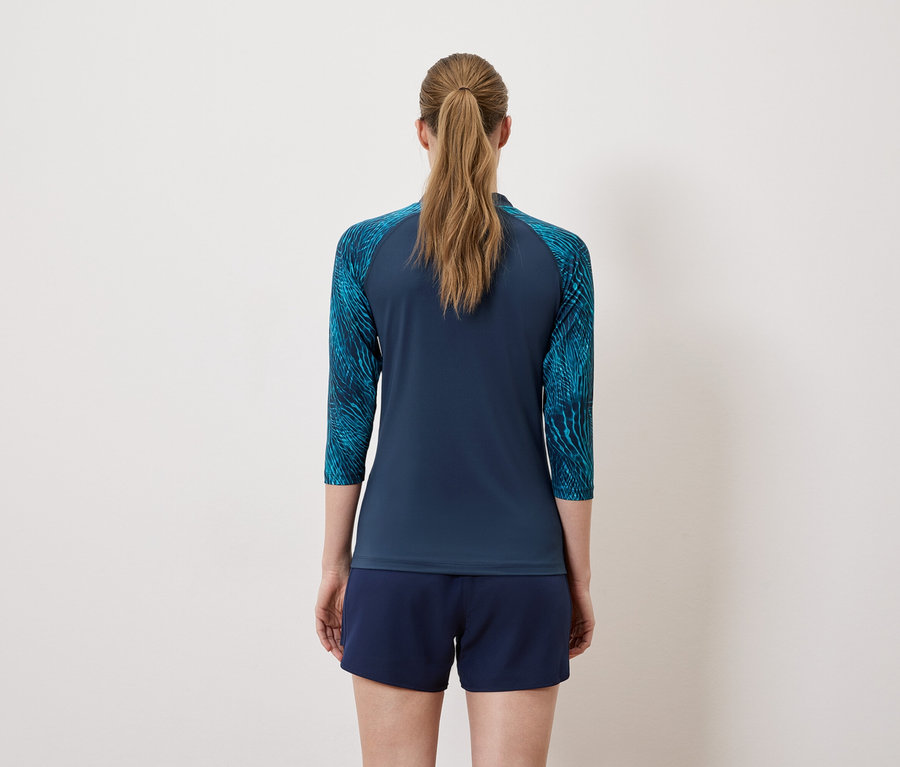 Rückansicht einer Frau mit Pferdeschwanz, die ein marineblaues, dreiviertelärmeliges Oberteil mit blaugrünen Ärmeln und marineblaue Shorts trägt.