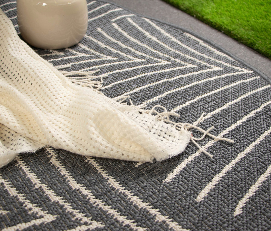 Ein cremefarbener Krug und eine geflochtene Decke liegen auf einem grauen Teppich mit weißem Zickzackmuster.