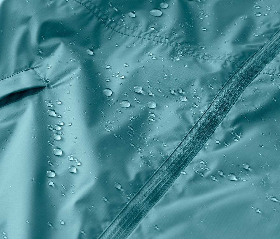 Nahaufnahme einer türkisfarbenen, wasserabweisenden Jacke mit Reißverschluss und einer Tasche, auf der Wassertropfen liegen.