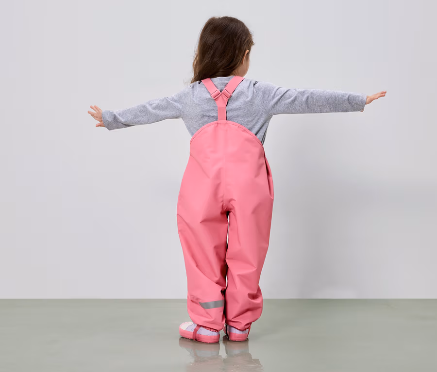 Mädchen von hinten in rosa Matschhose und grauem Oberteil mit ausgestreckten Armen vor einem hellen Hintergrund.