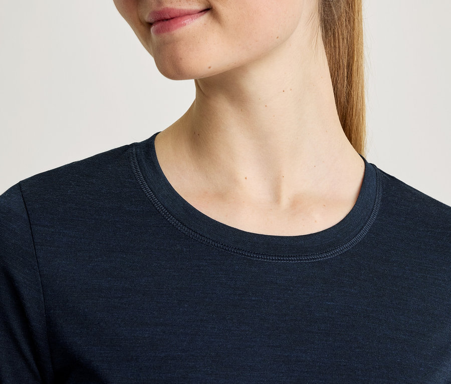 Frau trägt ein dunkelblaues Langarmshirt mit Rundhalsausschnitt. Ihr Haar ist zu einem Pferdeschwanz gebunden.